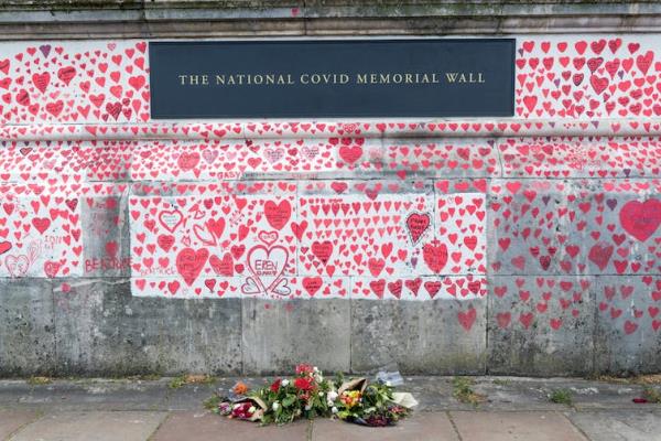 The UK's natio<em></em>nal covid memorial wall in London, a co<em></em>ncrete wall covered in illustrated red hearts, with a few bouquets of flowers at the ba<em></em>se.